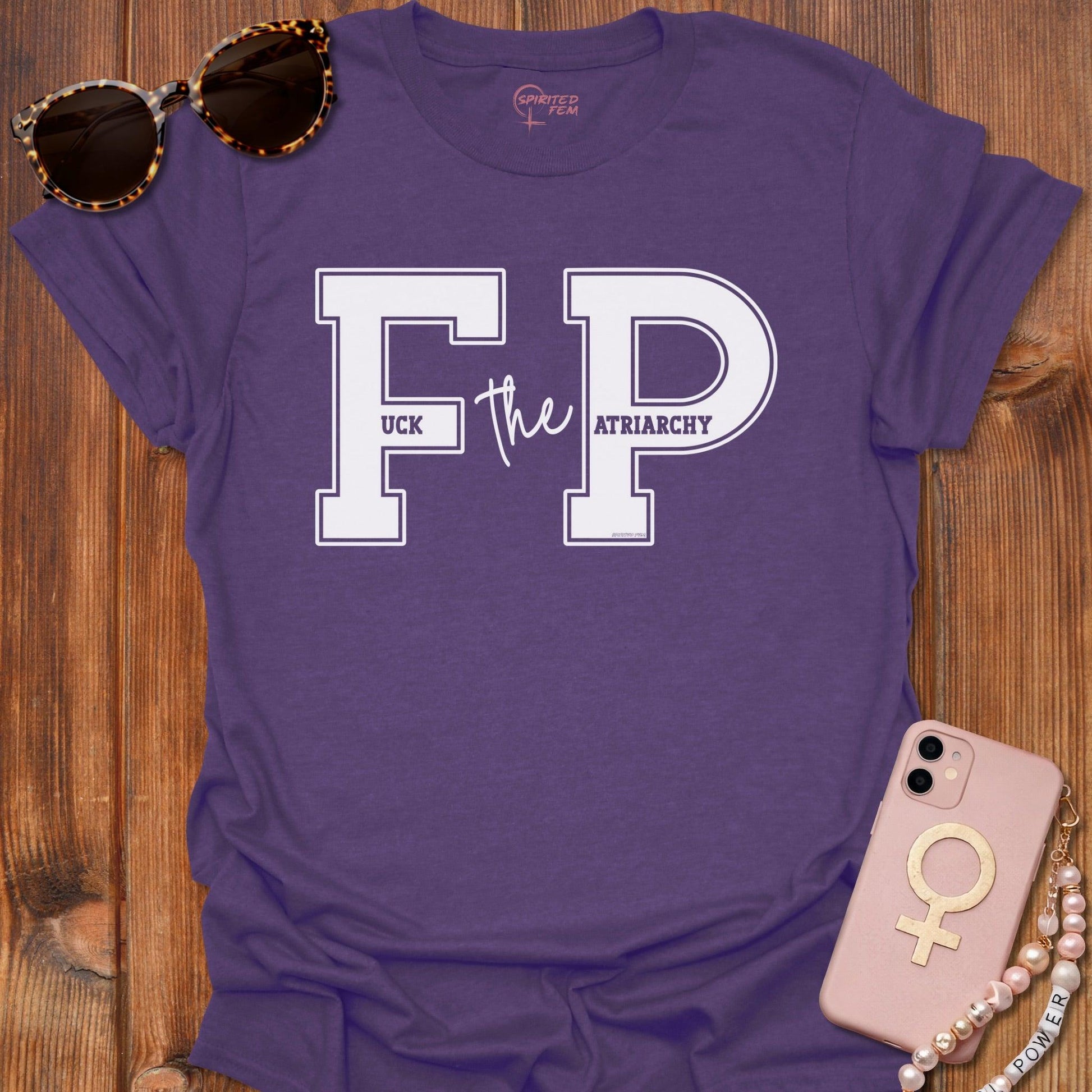 A purple graphic t-shirt laid flat on a wooden surface, styled with leopard-print sunglasses, a pink phone case, and a pearl bracelet. The t-shirt features a large white College Font reading 'FP' with 'the' in script and 'UCK ATRIARCHY' printed inside the letters. Altogether stating Fuck the Patriarchy
